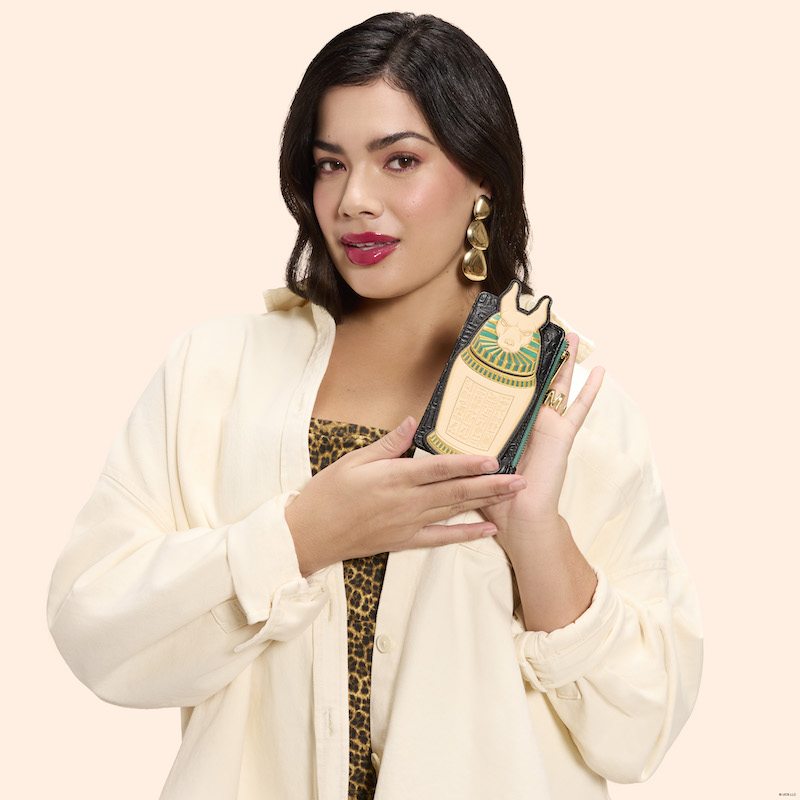 Woman wearing a cheetah-print bodysuit with a white jacket over it. She holds the Loungefly The Mummy large card holder in both hands. 